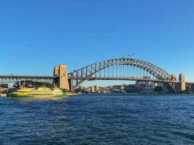 Sydney Harbour With The Metal Sydney Harbour Bridge With A Green And Gold Ferry Sailing In The Harbour.