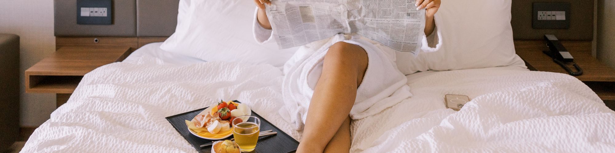 Person sitting on a bed with breakfast and reading a paper.